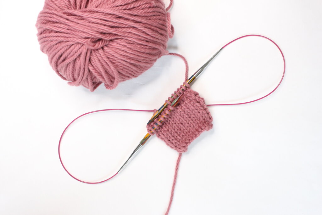 Deux boucles de câble formées de part et d'autre d'un petit tricot rose, sur des aiguilles circulaires interchangeables, pelote de laine rose posée en haut à gauche.