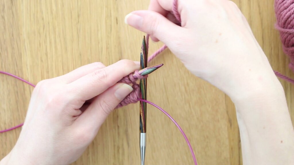 Premières mailles tricotées sur l'aiguille droite juste après avoir joint le tricot en rond, câble et pelote de laine rose en arrière-plan.