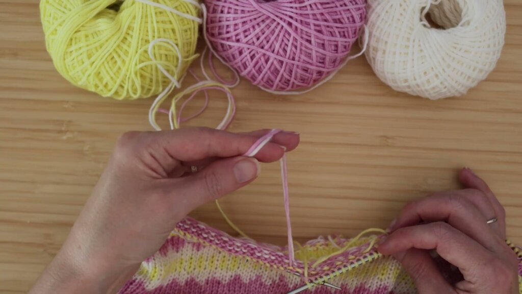 Trois pelotes jaune, rose et blanche reliées à un ouvrage en jacquard sur les aiguilles, pendant un changement de couleur